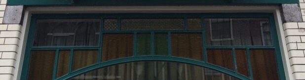 Detail met bovenlichten van winkelpui Lange Mare 53. Naast het gekleurde glas zijn drie ruitjes met ventilatieglas zichtbaar